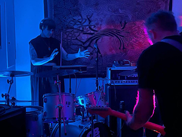 a blue room with two performers, one playing theremin and the other guitar. A drum kit stands between the two musicians
