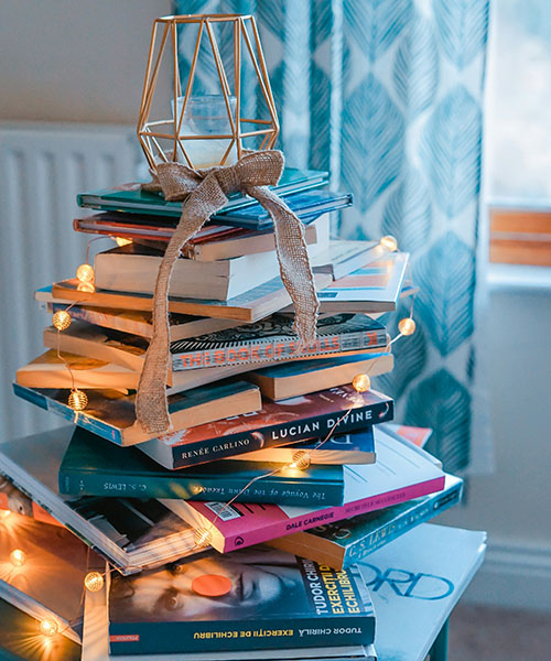 Pile of books stacked on top of each other with a candle encased in a brown-coloured structure with a bow and lights trailing down the books.